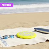 Yellow frisbee, sunglasses, and dark bottle rest on a white beach towel on the sand.