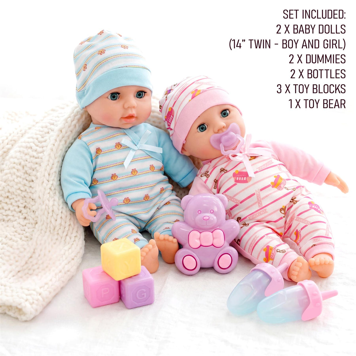 Two dolls sit with a bear toy, building blocks, bottles, and pacifiers arranged on a surface.