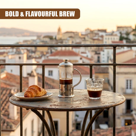 A metal cafetiere, a glass of coffee, and a croissant rest on a patterned outdoor table.
