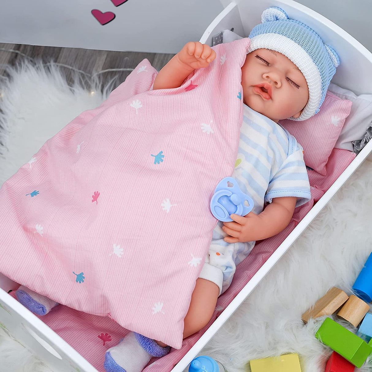 This image shows a baby doll lying in a pink baby bassinet with an attached blanket, wearing a blue and white knit cap, dressed in a onesie, surrounded by toys. The doll has a pacifier in its mouth, and there are colorful blocks in the corner of the photo. - UKBuyZone
