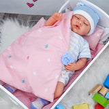 This image shows a baby doll lying in a pink baby bassinet with an attached blanket, wearing a blue and white knit cap, dressed in a onesie, surrounded by toys. The doll has a pacifier in its mouth, and there are colorful blocks in the corner of the photo. - UKBuyZone