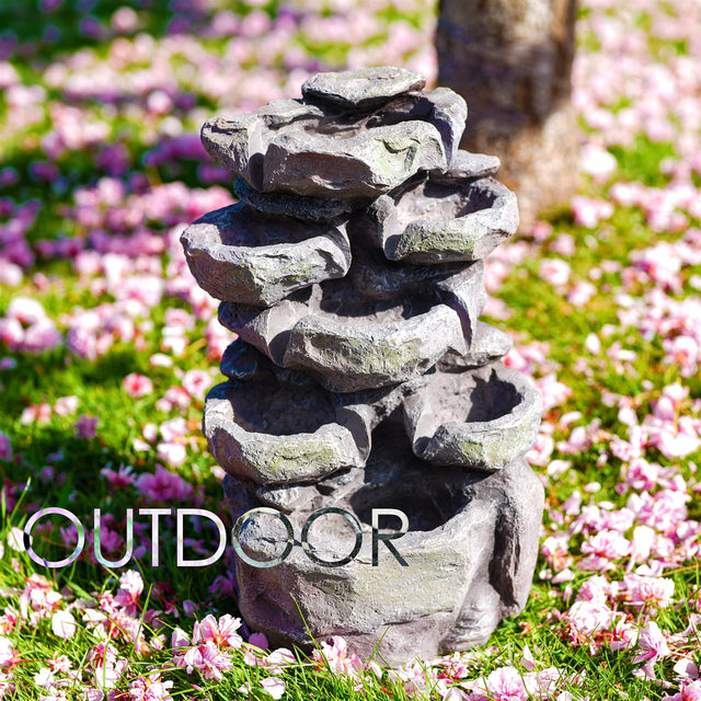 Multi-tiered stack of artificial gray rocks resting on vibrant green lawn and scattered pink blossoms.