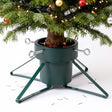 A green metal Christmas tree stand holding a decorated tree, featuring four adjustable bolts and a water reservoir base