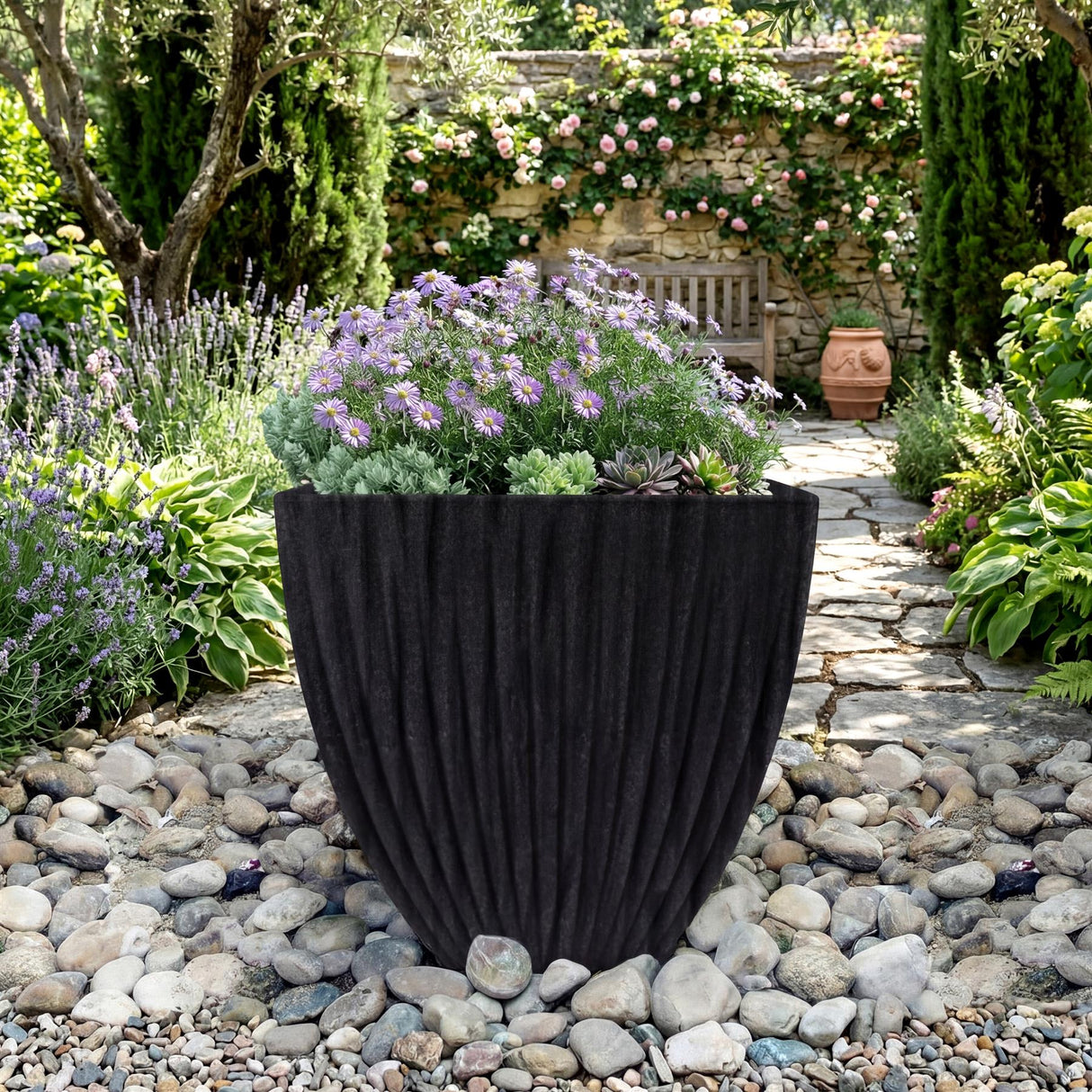 Large, pleated black planter containing lavender flowers, placed on light gray garden stones among greenery.