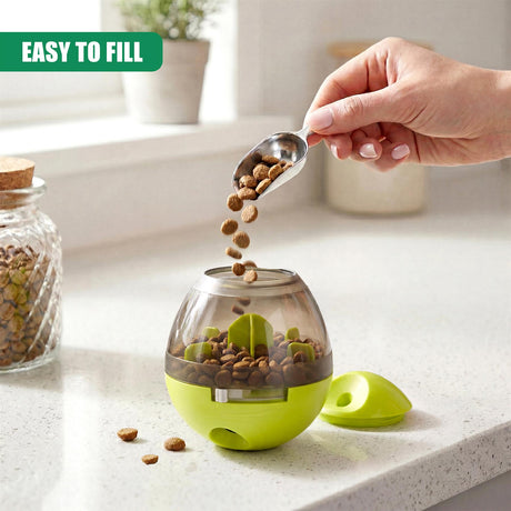 A hand pours dog treats from a spoonful into a green and clear plastic pet toy dispenser.