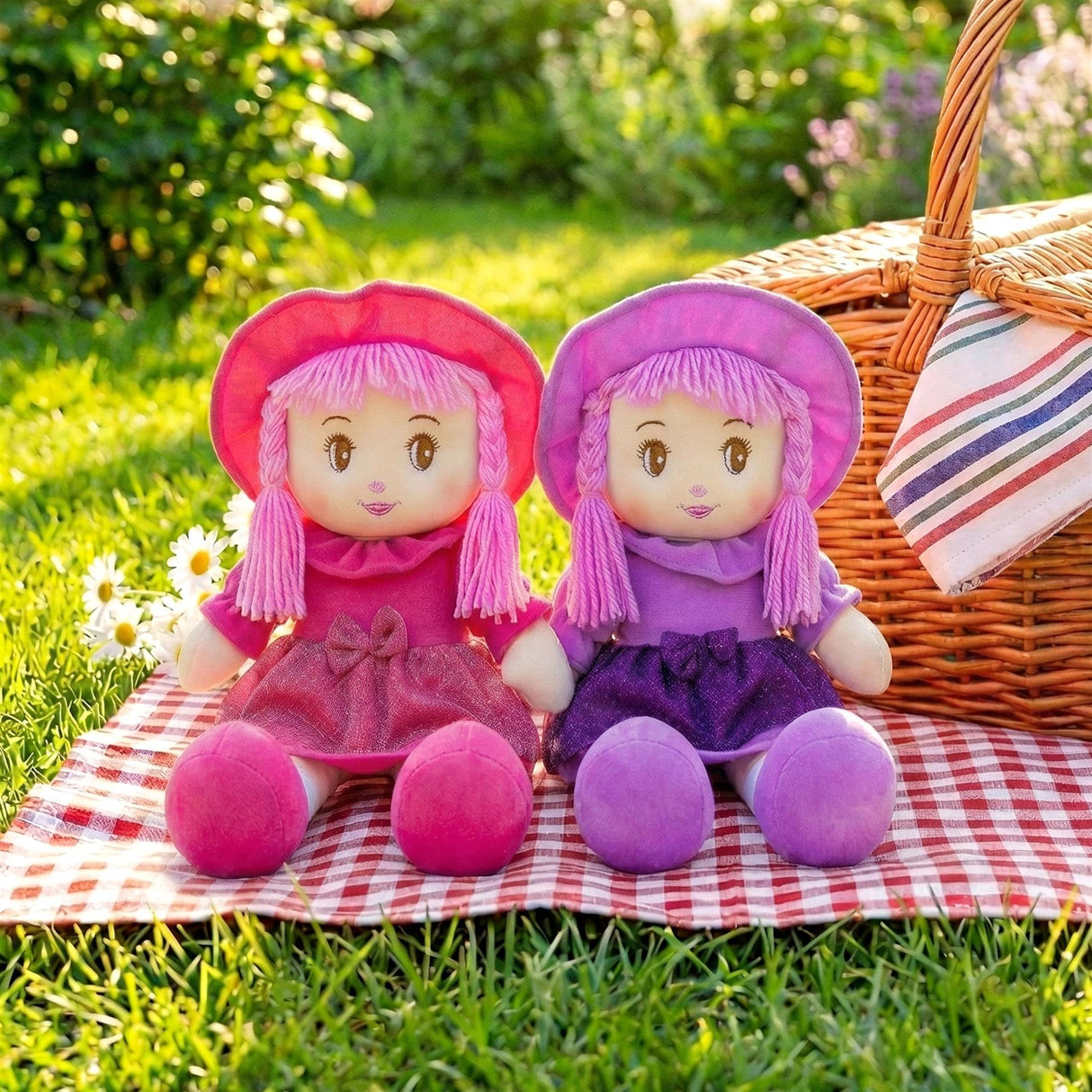 A pair of plush rag dolls in pink and purple sit on a checkered picnic cloth next to a woven basket.
