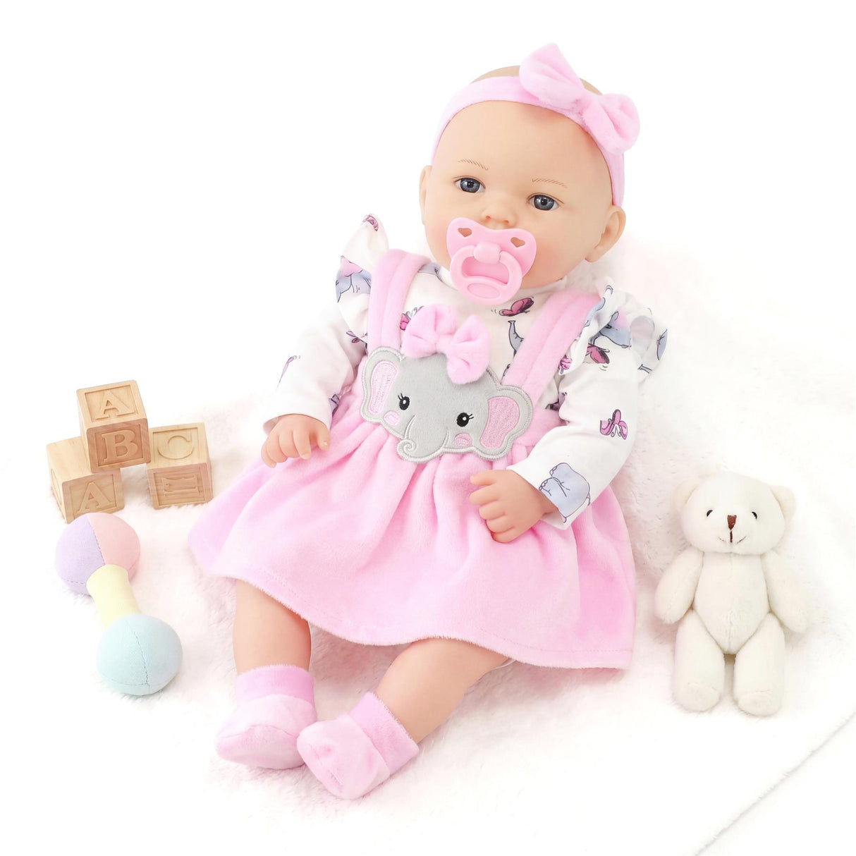 A doll in a pink dress lies with wooden blocks, a teddy bear, and a blue pacifier nearby.