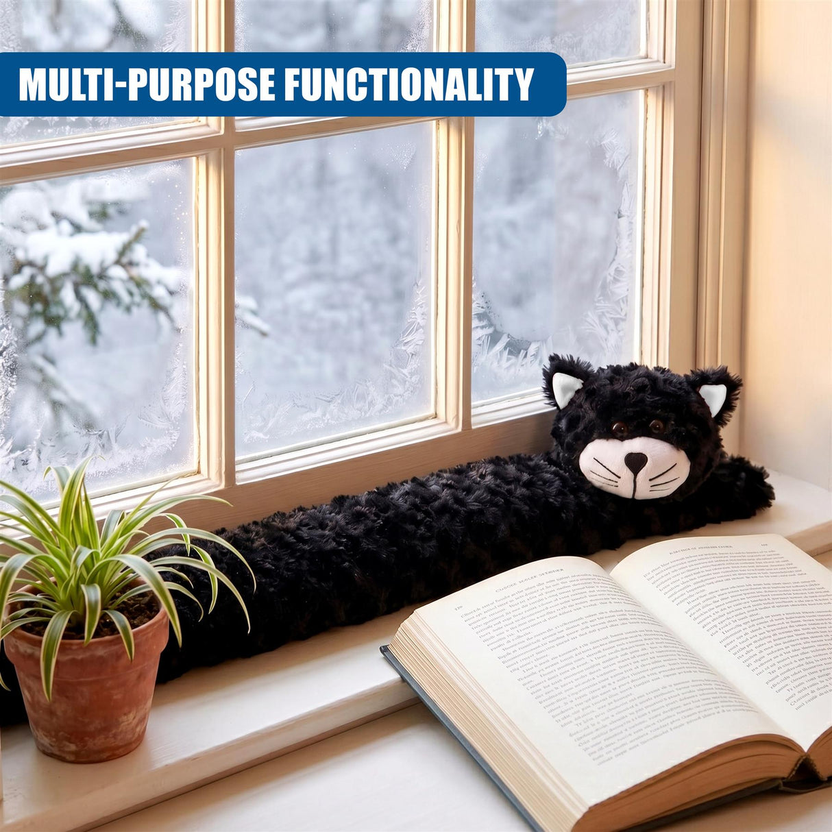 A black plush cat draught excluder rests on a window ledge with an open book and potted plant.