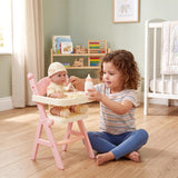 Girl feeding a baby doll at a pink doll high chair set with accessories.
