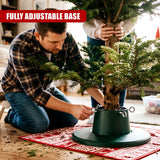 A man in a plaid shirt and jeans kneels on a red rug to adjust a green adjustable Christmas tree stand holding a real tree