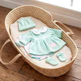 Mint green doll clothes set, including a dress, top, hat, and booties, arranged inside a woven basket.