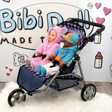 A blue, three-wheeled doll stroller holds two dolls dressed in pink and blue outfits.