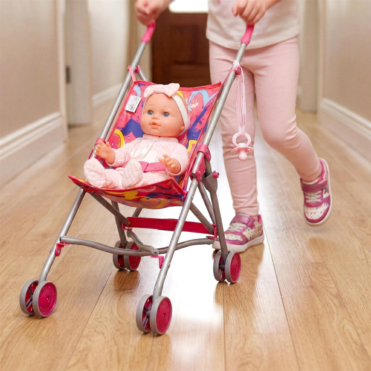 Pink stroller holding a doll is being pushed by a person down a wooden hallway floor.