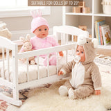 A doll in a pink bonnet sits in a crib next to a second doll wearing a bear-themed outfit.
