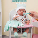 A reborn doll in mint and spotty clothes is sitting at a table while being fed by a hand.