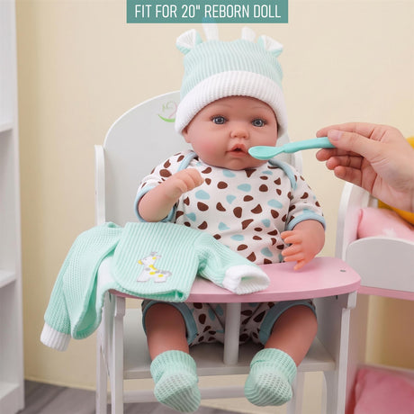 A reborn doll in mint and spotty clothes is sitting at a table while being fed by a hand.