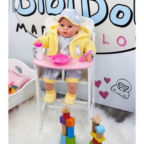 The image shows a young baby seated in a miniature high chair. The high chair is a wooden structure with a pink bowl and spoon on top, and it has a colorful design. In the background, there is a sign that reads "Bibliodog" and a banner with a pink ribbon at the bottom with text, although the specific text is not fully visible in this view of the image. The overall style of the image seems to be a casual indoor setting, likely a nursery or child's room. The high chair appears to be designed for … - UKBuyZone