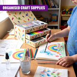 Art supplies and drawing papers are laid out on a table while a child draws a colorful rainbow design.