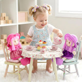 Pink-haired rag doll wearing a pink dress sits in a chair at a tea party setting.