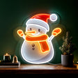 A GEEZY Christmas neon light shaped like a snowman wearing a red hat and orange scarf, displayed on a wooden shelf next to a