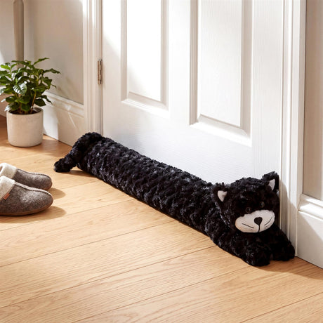 A plush black cat draught excluder rests against a white door on a light wooden floor.