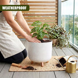 Hands planting a small green plant into a white ceramic pot supported by three visible wooden legs.