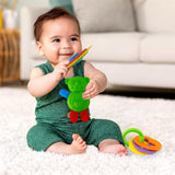 Baby holds colorful teether toys near a green reptile toy and stacked colorful rings.