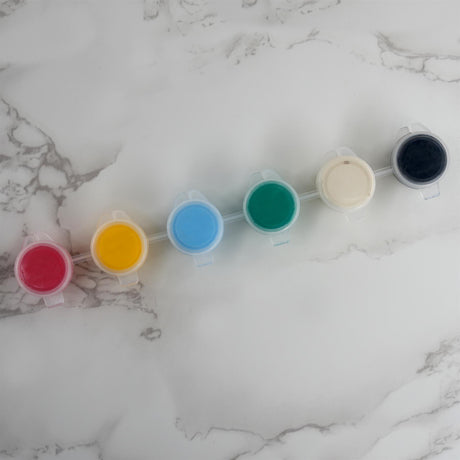 Six colored paint or material samples in clear containers displayed in a row on a white marble surface.