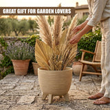 Beige pot with three legs filled with tall, dried decorative grasses and assorted foliage.