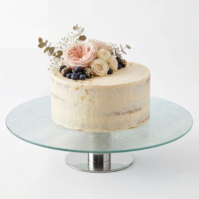 A clear glass, round cake stand with a central base, displaying a decorated cake with flowers and berries.