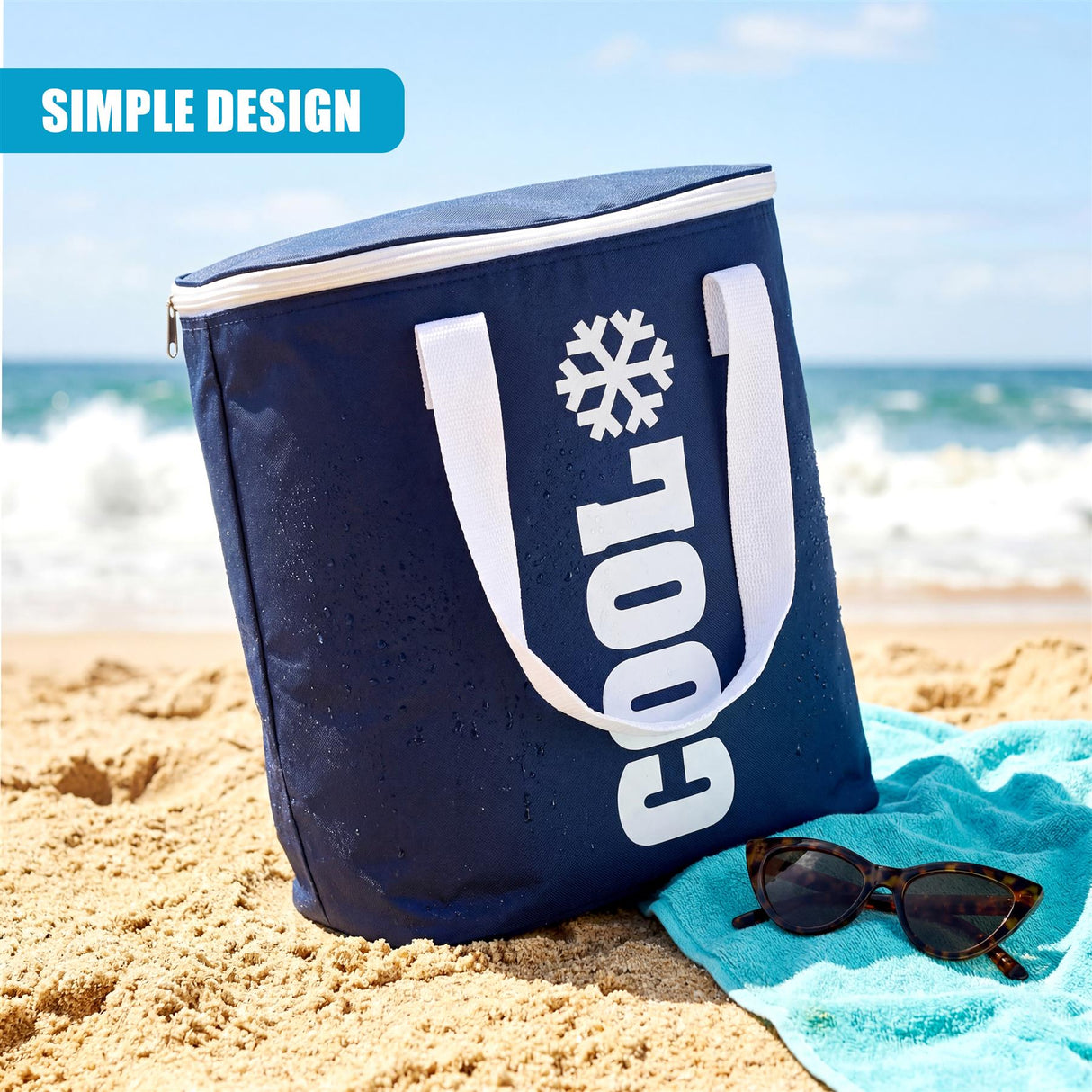 Navy cooler bag with white lettering and snowflake graphic sits on the sand near blue towel.