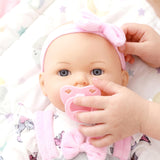 Lifelike baby doll with blue eyes and pink accessories holds a pacifier while resting.
