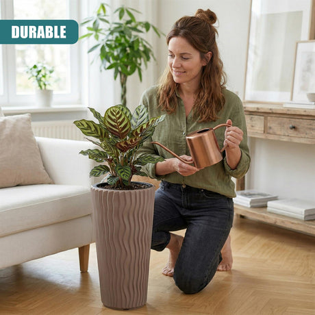 A tall, ribbed brown flower pot holds a leafy houseplant near a kneeling woman indoors.