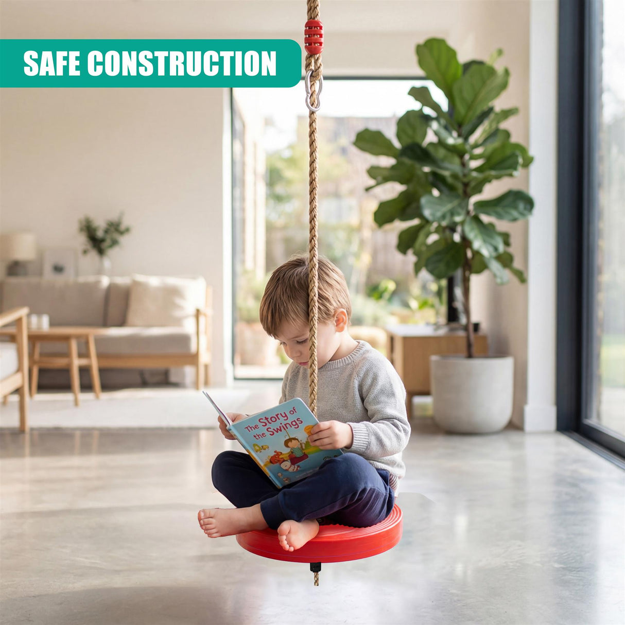 A young boy reads a book while sitting on a red swing seat suspended by ropes.