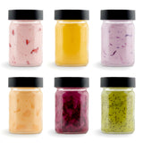 Six small glass jars with black lids holding various colored powdered seasonings.