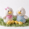 Two white plush chicks and yellow decorative eggs rest in a bed of straw.