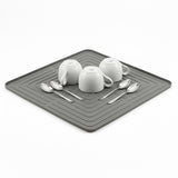 White dishes and metal spoons are displayed on a dark gray rectangular drying mat.