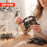 A woman holds a black Christmas light reel storage device over a wooden table, with fairy lights and spare reels nearby