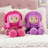 Pink and purple rag dolls with hoods and pigtails rest on a soft, cushioned surface.