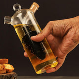 A hand holding a clear glass GEEZY 2 in 1 Oil and Vinegar Dispenser, filled with golden oil and featuring a black silicone pourer inside. The dispenser has a cork stopper at the top, with a wooden surface and slices of bread in the foreground. Perfect for serving oils and vinegars decoratively, enhancing your dining experience. Available at ukbuyzone.