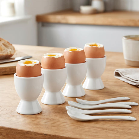 A set of four white ceramic egg cups holding boiled eggs on a wooden table, accompanied by matching spoons