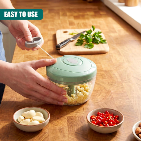 Ingredients like garlic, peppers, and herbs surround a manual food chopper on a wooden counter.