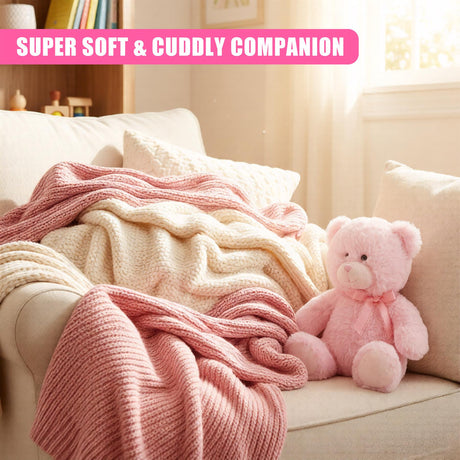 A plush, pink teddy bear sits on a cream-colored couch, surrounded by a soft, cream-colored blanket and a pink, chunky throw blanket. The teddy bear is adorned with a pink bow and has a sweet, smiling face. The background is a warm, sunny room with a window and a bookshelf in the background. - UKBuyZone