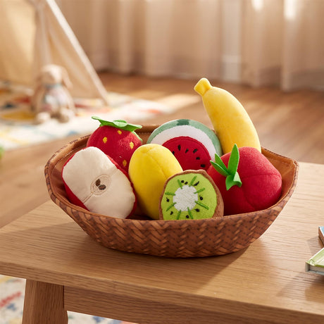 A brown woven fabric basket filled with seven soft plush fruits including a banana, strawberry, watermelon, kiwi, apple and