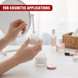 A hand scoops white cream from a small glass jar using a miniature metal applicator.
