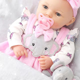 A doll wears a pink and white patterned outfit, holding a plush gray elephant toy.