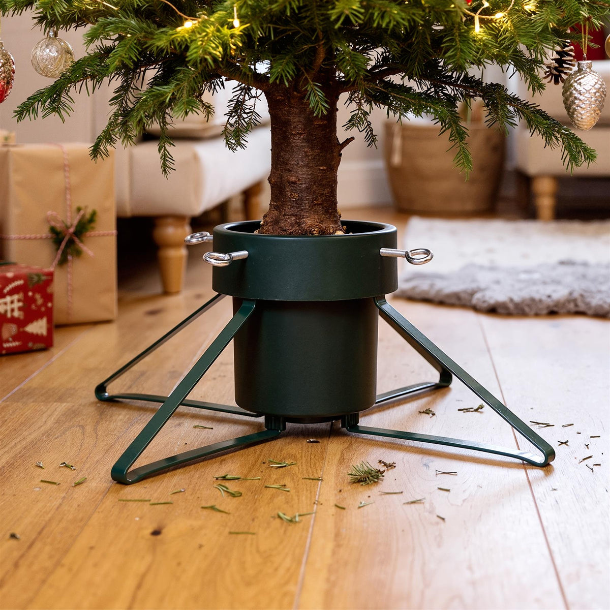 A green metal Christmas tree stand holding a decorated tree on a wooden floor with scattered pine needles and wrapped gifts