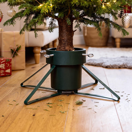A green metal Christmas tree stand holding a decorated tree on a wooden floor with scattered pine needles and wrapped gifts
