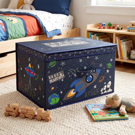 A large navy blue space-themed storage box with a lid, decorated with rockets, planets and stars, sits on a cream rug