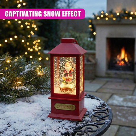 An illuminated red lantern displaying a Christmas scene on a snowy outdoor wrought-iron table.